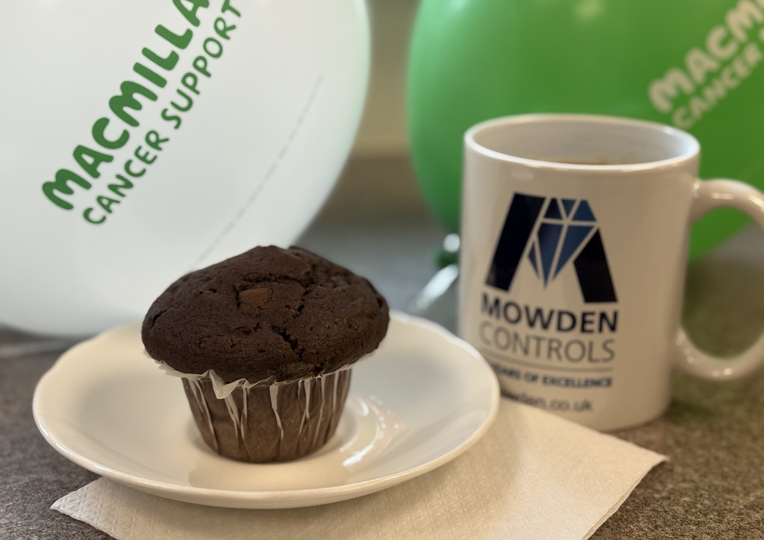 A cup of coffee and a chocolate muffin with a background of MacMillan cancer support balloons.